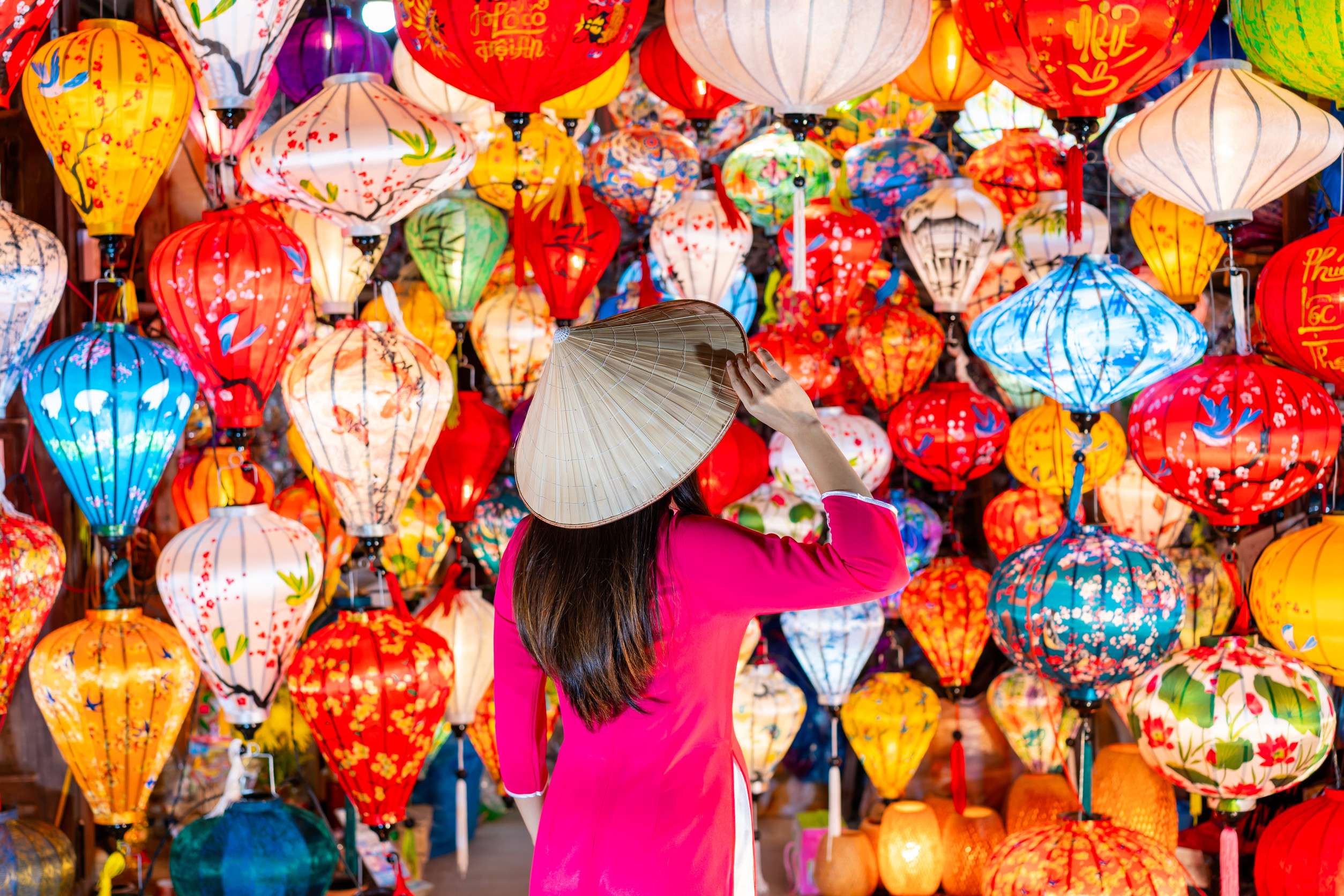 Young female tourist in Vietnamese traditional dress looking at paper ornamental lanterns in Hoi An Young female tourist in Vietnamese traditional dress looking at paper ornamental lanterns in Hoi An
